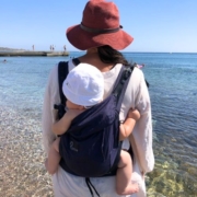 Kundin mit dunkelblauer Leolin Mini Trage und Baby auf dem Rücken vor einem Meer stehend